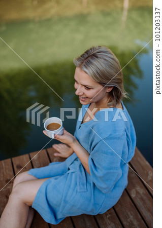 Happy young woman sitting by pond near cottege and enjoying cup of morning coffee on summer vacation in mountains. High angle view. 101203137