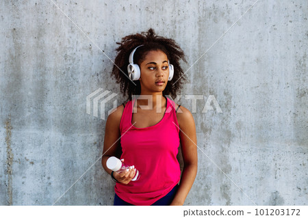 Young multiracial girl in sportswear resting after jogging in city, drinking water from sustainable bottle and listening music in headphones. 101203172