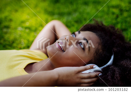 Multiracial girl lying down in grass and enjoying music in headphones, side view. 101203182