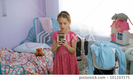 Little girl in pink dress grimacing while taking selfies on her smart phone in messy room 101203575