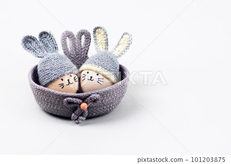 Two Easter eggs with funny faces and gray crocheted hats with bunny ears in a decorative basket on a gray background. Happy Easter concept. Greeting card. Two Easter eggs with funny faces and gray crocheted hats with bunny ears in a decorative basket on a gray background. Happy Easter concept. Greeting card. 101203875