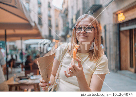 Happy woman eating traditional spanish delicious churros, a fried pastry with chocolate near a city cafe in Valence, Spain 101204074