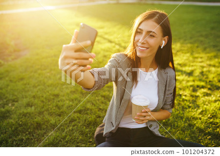 Time to take selfie! Image of beautiful stylish woman sitting on green grass. 101204372