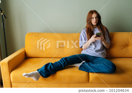 Portrait of serious thoughtful young woman holding smartphone, sitting on couch at home, thinking over online chat text message, looking away. Redhead female using mobile phone, getting bad news. 101204634