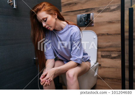 Portrait of tired young woman in casual clothes sitting on toilet bowl at home bathroom with modern interior. Bored exhausted redhead female sitting in restroom after party feeling sleepy. Portrait of tired young woman in casual clothes sitting on toilet bowl at home bathroom with modern interior. Bored exhausted redhead female sitting in restroom after party feeling sleepy. 101204680