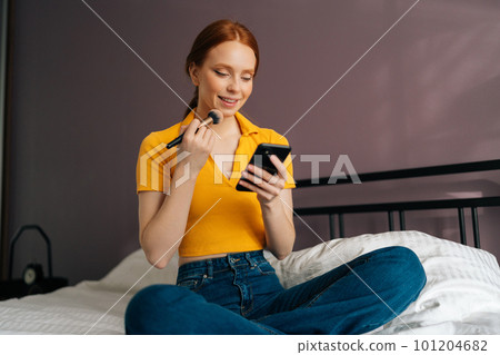 Vertical portrait of smiling redhead young woman applying cosmetic powder with big professional makeup brush and using smartphone. Pretty female using blusher with brush and reading social media. Vertical portrait of smiling redhead young woman applying cosmetic powder with big professional makeup brush and using smartphone. Pretty female using blusher with brush and reading social media. 101204682