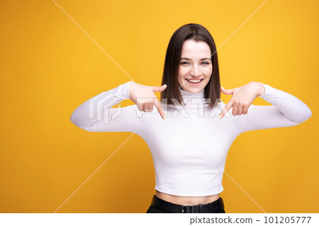 The woman smiles and points down. Portrait of a girl in a white blouse on a yellow background in the studio. The woman smiles and points down. Portrait of a girl in a white blouse on a yellow background in the studio. 101205777