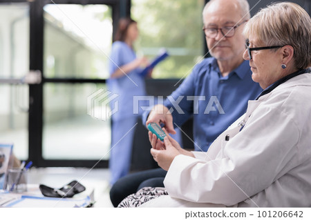 Senior medic taking patient insulin level test using glucometer in hospital waiting area, doing diabetes examination to measure glucose. Elderly man having checkup visit consultation 101206642
