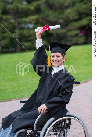 Caucasian woman in a wheelchair in a graduate costume rejoices at receiving a diploma.  101210117