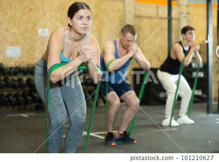 Motivated young woman practicing exercises with stretch rope sitting near other people in gym Motivated young woman practicing exercises with stretch rope sitting near other people in gym 101210627