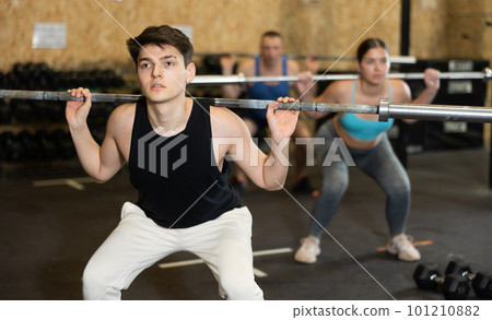 Motivated young man practicing exercises with rod near other people in gym Motivated young man practicing exercises with rod near other people in gym 101210882