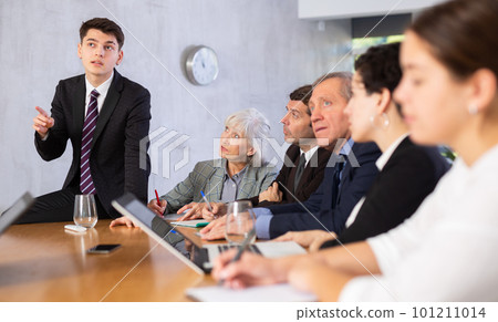 Young male leaning on table and sharing ideas while his colleagues using laptops and making notes in meeting room Young male leaning on table and sharing ideas while his colleagues using laptops and making notes in meeting room 101211014