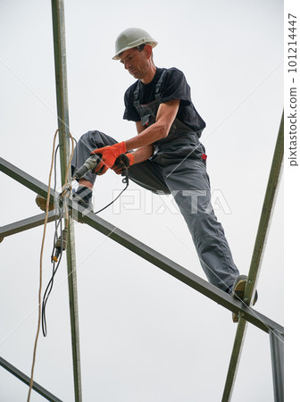 Full length of man in safety helmet and work overalls standing on metal rail and using electric drill while installing support structure for photovoltaic solar panels. Isolated on white background. Full length of man in safety helmet and work overalls standing on metal rail and using electric drill while installing support structure for photovoltaic solar panels. Isolated on white background. 101214447