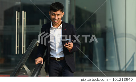 Smiling young businessman holding smart phone walking up stairs and looking away. Urban lifestyle and technology concept 101218940