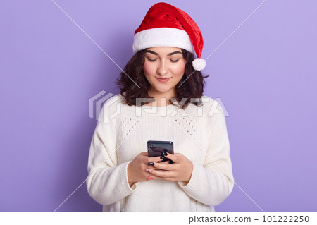 Image of pleasant cute dark haired young lady having peaceful facial expression, holding mobile phone in both hands, chatting, using social networking sites. People and new technologies concept. 101222250