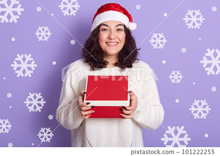 Picture of cheerful peaceful young woman wearing red santa claus hat and white sweater, holding red box in both hands, getting christmas presents, standing isolated over lilac background in studio. Picture of cheerful peaceful young woman wearing red santa claus hat and white sweater, holding red box in both hands, getting christmas presents, standing isolated over lilac background in studio. 101222255