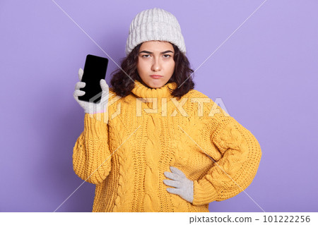 Close up portrait of curly haired disappointed woman, having frowned facial expression, looking directly at camera, wearing yellow sweater and white hat, holding smartphone, putting hand on waist. 101222256
