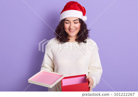 Indoor studio shot of happy beautiful female wearing red santa claus hat and white sweater, standing isolated over lilac background, holding box, opening to see present for new year. Holidays concept. 101222267