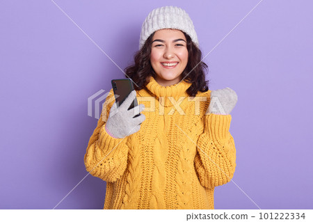 Picture of energetic beautiful female with black curly hair, clenching fist, holding smartphone, smiling sincerely, having pleasant facial expression, being in high spirits. Modern devices concept. 101222334
