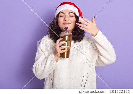 Photo of delighted peaceful young curly haired girl closing her eyes, feeling smell of coffee from thermal cup, enjoying hot drink, raising one hand, wearing hat and white sweater. Atmosphere concept. 101222335