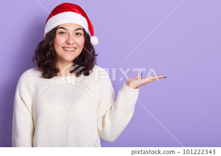 Indoor studio shot of pleasant good looking brunette raising hand, making gesture, looking directly at camera, wearing new year hat and white sweater. Copyspace for advertisement. Christmas concept. 101222343