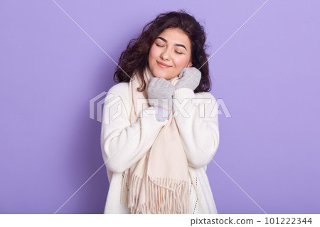Pleasant positive beautiful female closing eyes, smiling sincerely, spending time alone, wearing white scarf, sweater and gloves, standing isolated over lilac background in studio. Winter concept. 101222344