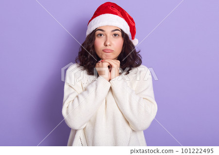 Cute woman with dark wavy hair posing dresses warm whiete sweater and Santa Claus cap, female looking at camera with pouty lips and keeps her fisnts under chin, isolated over purple studio background. 101222495
