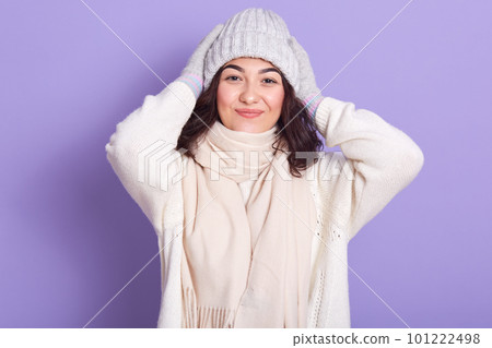 Image of curly haired beautiful pleasant young lady putting her hands on head, having positive facial expression, looking directly at camera, wearing white sweater and scarf, grey hat and gloves. 101222498