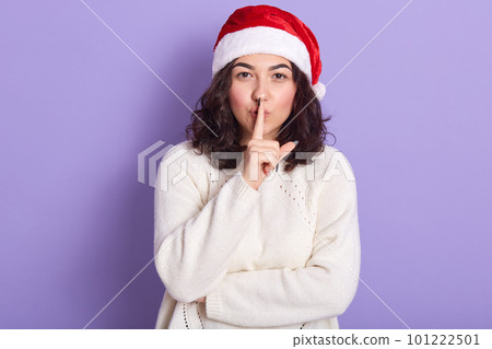 Mysterious good looking brunette with curly black hair standing isolated over lilac background in studio, making gesture, putting finger close to lips, making sh sound. People and gestures concept. 101222501