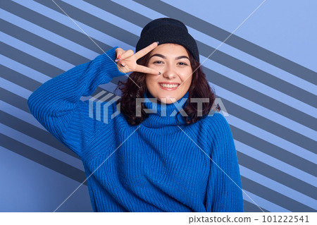 Adorable cheerful good looking model with dark curly hair smiling sincerely, looking directly at camera, making gesture, showing peace sign, wearing blue hat and sweater, having stripes aback. Adorable cheerful good looking model with dark curly hair smiling sincerely, looking directly at camera, making gesture, showing peace sign, wearing blue hat and sweater, having stripes aback. 101222541