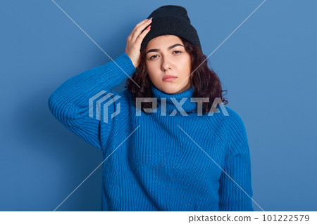 Portrait of thoughtful upset disappointed young lady with black curly hair, touching her head with hand, feeling unwell, having headache, standing isolated over phantom blue background in studio. Portrait of thoughtful upset disappointed young lady with black curly hair, touching her head with hand, feeling unwell, having headache, standing isolated over phantom blue background in studio. 101222579