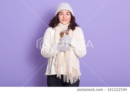 Beautiful girl basking her hands with thermo mug, enjoying to drink hot tea or coffee in cold winter weather, dresses warm sweater, scarf, gloves and cap, posing isolated over purple background. Beautiful girl basking her hands with thermo mug, enjoying to drink hot tea or coffee in cold winter weather, dresses warm sweater, scarf, gloves and cap, posing isolated over purple background. 101222594