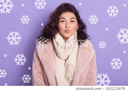 Indoor shot of stylish lady wearing pale pink oversize eco fur coat blowing kiss. Portrait of wavy haired woman posing over lilackstudio wall with snowflakes on background. People life style concept. 101222679