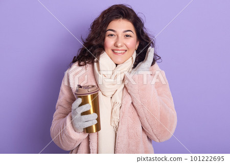 Close up portrait of happy girl with thermo mug of coffee or tea, looking ar camera and smiling, wearing eco fur coat, keeps palm raised, posing isolated over purple studio background. Winter concept. 101222695