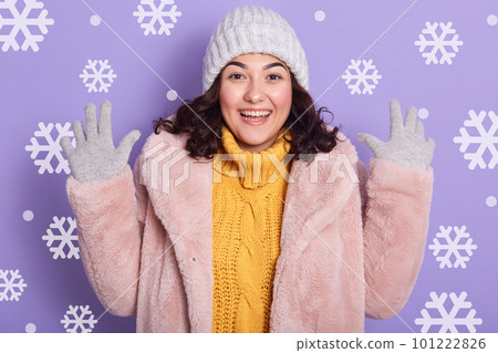 Image of woman wearing stylish eco fur coat, yellow sweater and cap, looking at camera and keeping hands up in studio with snowflakes on background. Female looks excited and expresses happyness. Image of woman wearing stylish eco fur coat, yellow sweater and cap, looking at camera and keeping hands up in studio with snowflakes on background. Female looks excited and expresses happyness. 101222826