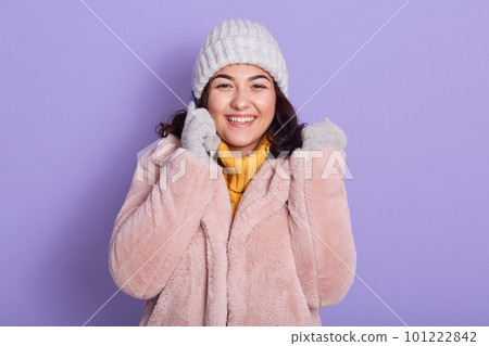 Cheerful pretty talking via phone, listening fresh news, laughing, wear pink fur coat warm yellow knitted pullover, cap and mittens, isolated over lilac color background. 101222842