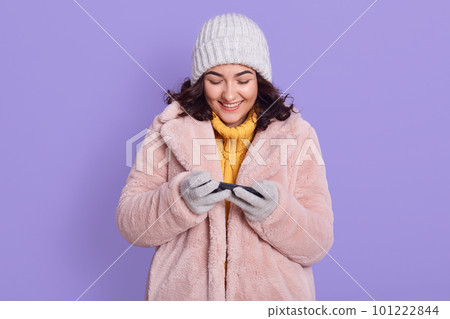 Young smiling female wearing winter fluffy coat and cap, standing with smart phone in hands, chatting or using app, isolated over lilac background, girl looking at screen of her device. Young smiling female wearing winter fluffy coat and cap, standing with smart phone in hands, chatting or using app, isolated over lilac background, girl looking at screen of her device. 101222844