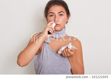 Sick woman holding wipe in her hands and using nasal spray, posing isolated over white background, wearing casual clothing, feels unwell, girl looking at camera with tortured facial expression. 101222845