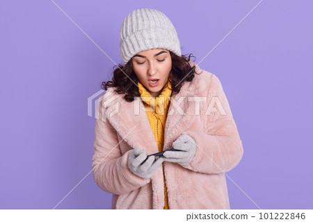 Astonished startled cute dark haired woman in fur coat just won challenge in awesome game, holds smart phone horizonally, plays racing, stares at display amazed, posing isolated over lilac background. Astonished startled cute dark haired woman in fur coat just won challenge in awesome game, holds smart phone horizonally, plays racing, stares at display amazed, posing isolated over lilac background. 101222846