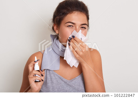 Brubette unhealthy Caucasian woman with napkin sneezing, holding nasal spray in hands to help herself, experiences allergy symptoms or caughting. Rhinitis, cold, sickness and allergy concept. 101222847