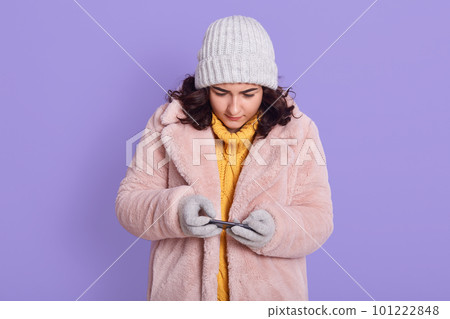 Young beautiful concentrated woman posing isolated over lilac wall background, playing games by mobile phone, looking at display, wearing faux fur coat, cap and mittens. Young beautiful concentrated woman posing isolated over lilac wall background, playing games by mobile phone, looking at display, wearing faux fur coat, cap and mittens. 101222848