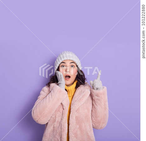 Astonished young woman wearing faux fur and cap pointing index finger up on copy space and keeps other palm on cheek, isolated over lilac background. People sincere emotions, lifestyle concept. 101222880