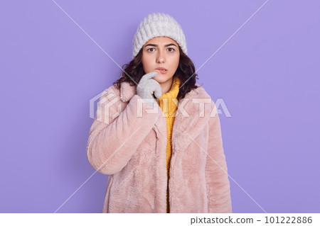 Pensive good looking young woman with dark hair looking thoughtfully at camera, dressed in fashionable pink coat and cap, looking concentrated, poses against blue background, keeping finger on lip. 101222886