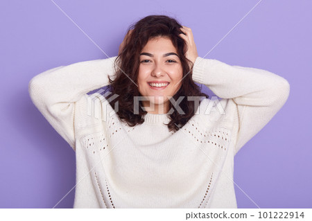 Happy smiling female wearing white sweater looking directly at camera with excited facial expression, keeping hands on head, smiling happily against lilac background, hears excellent news. 101222914
