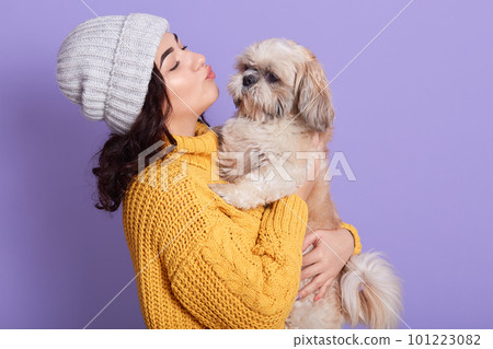 Portrait of beautiful girl keeping pretty Pekingese and wants kissing him, side view of female looks at her favorite puppy, lady in warm clothing, standing isolated over lilac background. Portrait of beautiful girl keeping pretty Pekingese and wants kissing him, side view of female looks at her favorite puppy, lady in warm clothing, standing isolated over lilac background. 101223082