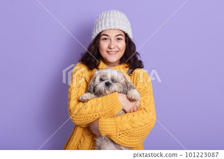 Female hugging her puppy and looking directly at camera with satisfied facial expression, brunette girl with wavy hair dresses white cap and yellow knitted warm jumper, isolated over lilac background. Female hugging her puppy and looking directly at camera with satisfied facial expression, brunette girl with wavy hair dresses white cap and yellow knitted warm jumper, isolated over lilac background. 101223087