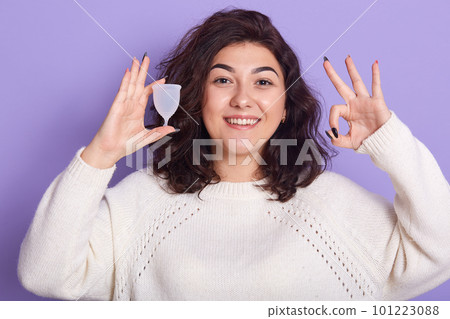 Close up of portrait of young woman holding silicon menstrual cup isolated over lilac background in studio, showing ok sign, feelsgood during her period. Modern eco friendly female hygiene concept. Close up of portrait of young woman holding silicon menstrual cup isolated over lilac background in studio, showing ok sign, feelsgood during her period. Modern eco friendly female hygiene concept. 101223088