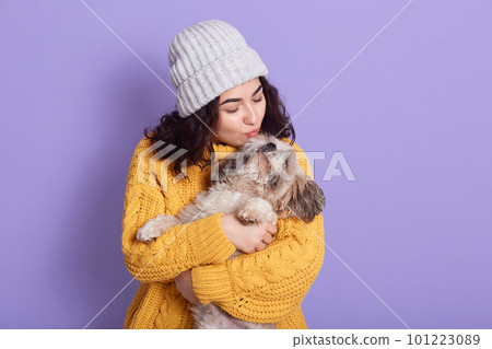 Beautiful young woman with funny Pekingese dog on lilac background, girl looks at her pet and wants to kiss puppy, lady in warm clothing and cap against lilac wall. Beautiful young woman with funny Pekingese dog on lilac background, girl looks at her pet and wants to kiss puppy, lady in warm clothing and cap against lilac wall. 101223089