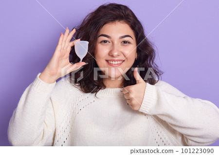 Close up portrait of woman holding menstrual cup isolated over purple studio background, happy female wearing white sweater showing thumb up, having period, dislike to use napkins or pampons. Close up portrait of woman holding menstrual cup isolated over purple studio background, happy female wearing white sweater showing thumb up, having period, dislike to use napkins or pampons. 101223090