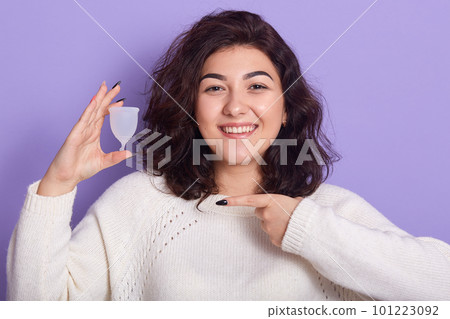 Horizontal shot of brunette female holding in hand plastic menstrual cup isolated over purple studio background, smiling woman pointing at her personal hygiene product. PMS and period concept. Horizontal shot of brunette female holding in hand plastic menstrual cup isolated over purple studio background, smiling woman pointing at her personal hygiene product. PMS and period concept. 101223092
